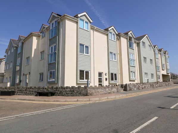A row of modern multi-story residential buildings with a stone wall in front, a clear sky, and a road in the foreground. - Maelog, 14 Tides Reach (Rhosneigr)
