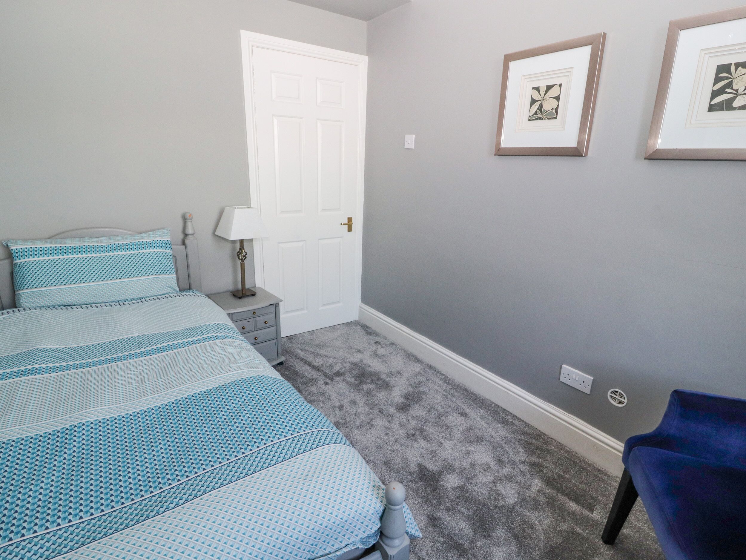 A bedroom with a bed, a nightstand, a lamp, a door, and two framed pictures on the wall.