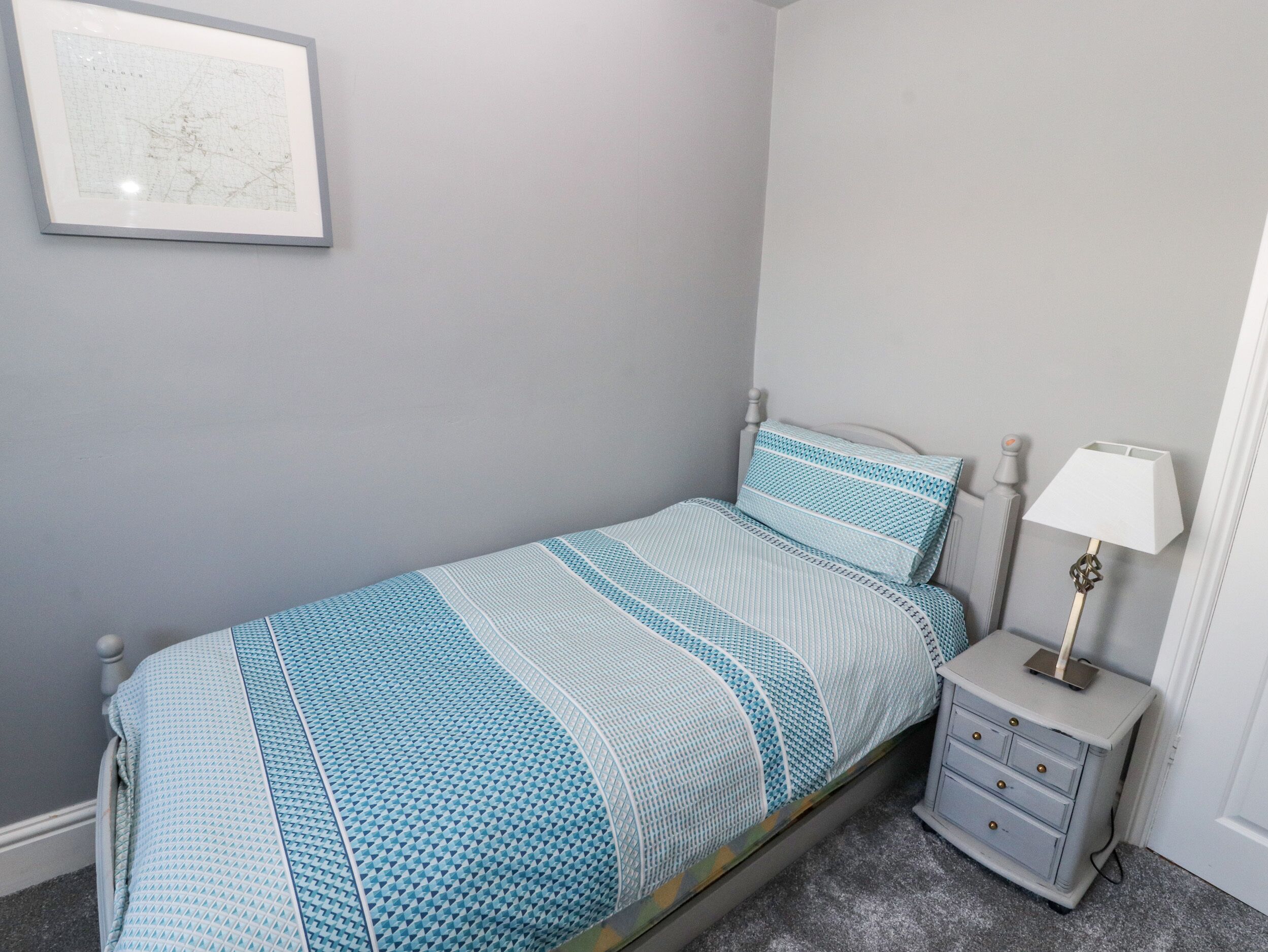A bedroom with a single bed, a nightstand with a lamp, and a framed map on the wall.
