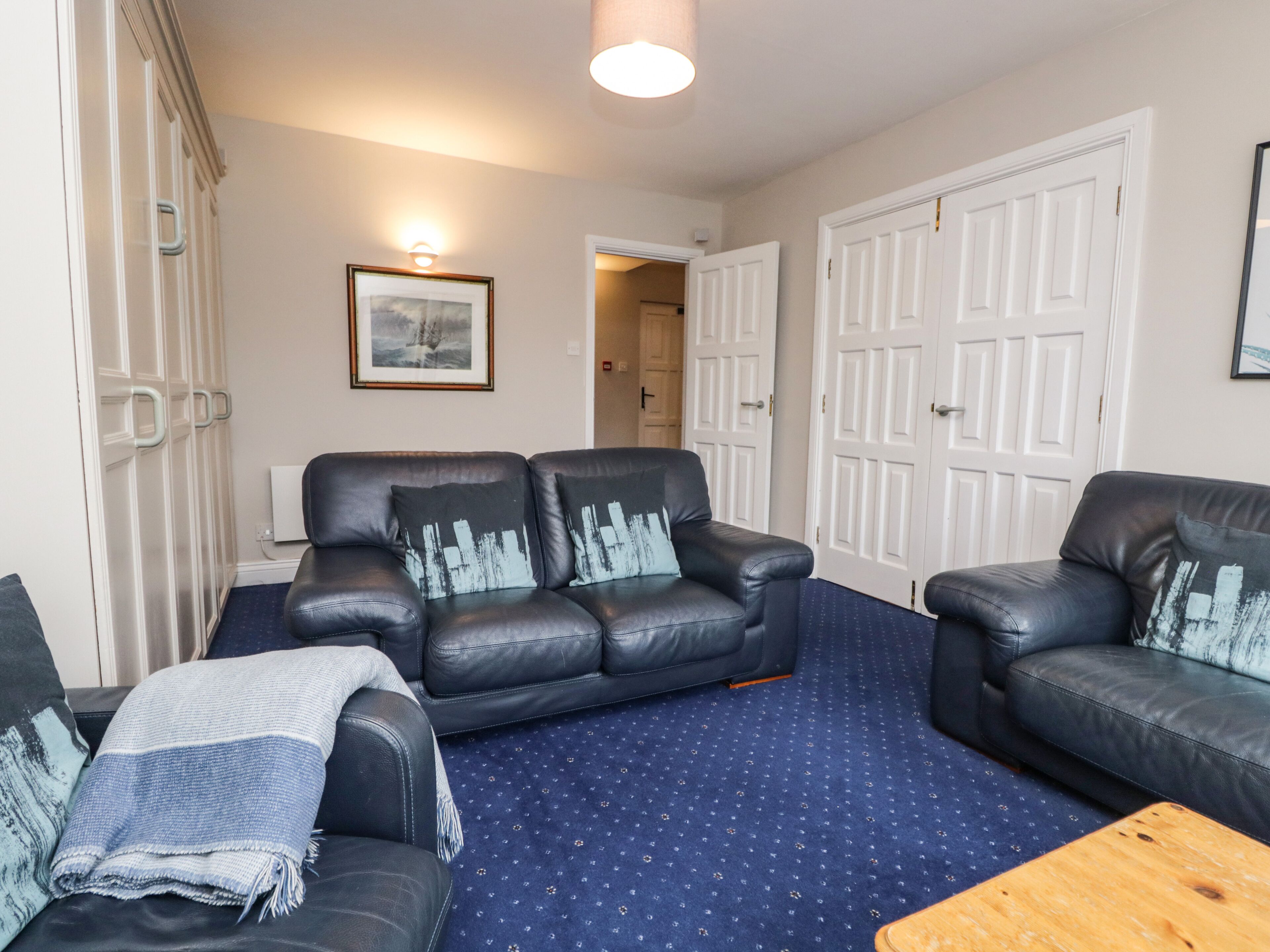A living room with a blue carpet, two black leather sofas with patterned cushions, a wooden coffee table, and white doors in the background.