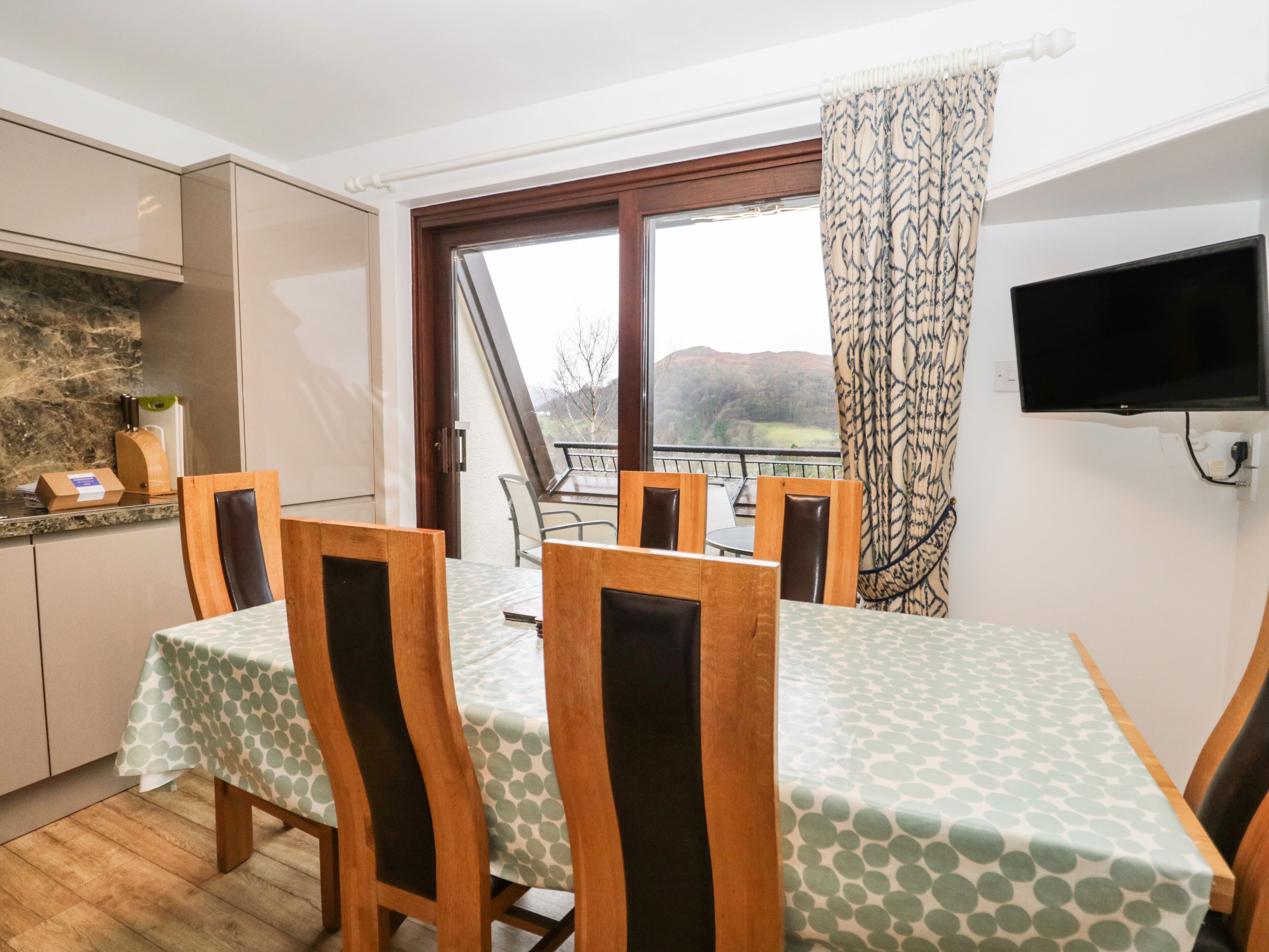Ein Essbereich mit Tisch für vier Personen, Holzsesseln, einem Fenster mit Blick auf die Landschaft und einem an der Wand befestigten Flachbildfernseher.