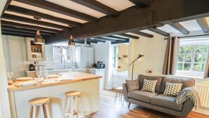 A modern kitchen with a central island, wooden stools, and a grey sofa in a living area with a large window. - 4 Masons Court (Stratford-upon-Avon)