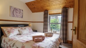 A bedroom with a wooden ceiling, a large bed with floral bedding, a nightstand, a window with curtains, and a door. - Cae Tanws Bach (Dolgellau)