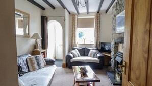 A cozy living room with a sofa, a coffee table, and a television. There is a window with curtains, a lamp, and a small table with decorative items. - Cae Tanws Bach (Dolgellau)