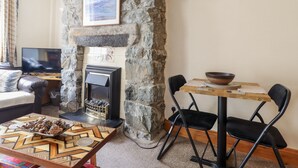 A cozy living room with a stone fireplace, a television, and a dining area with chairs and a table. - Cae Tanws Bach (Dolgellau)