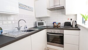 A compact kitchen with white cabinets, a stainless steel oven, a microwave, a toaster, and a dish rack. - Sandy Shell Cottage (Conwy)