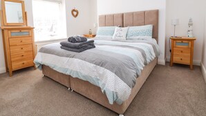 A bedroom with a large bed, wooden nightstands, a dresser, a mirror, and a window with blinds. - Sandy Shell Cottage (Conwy)
