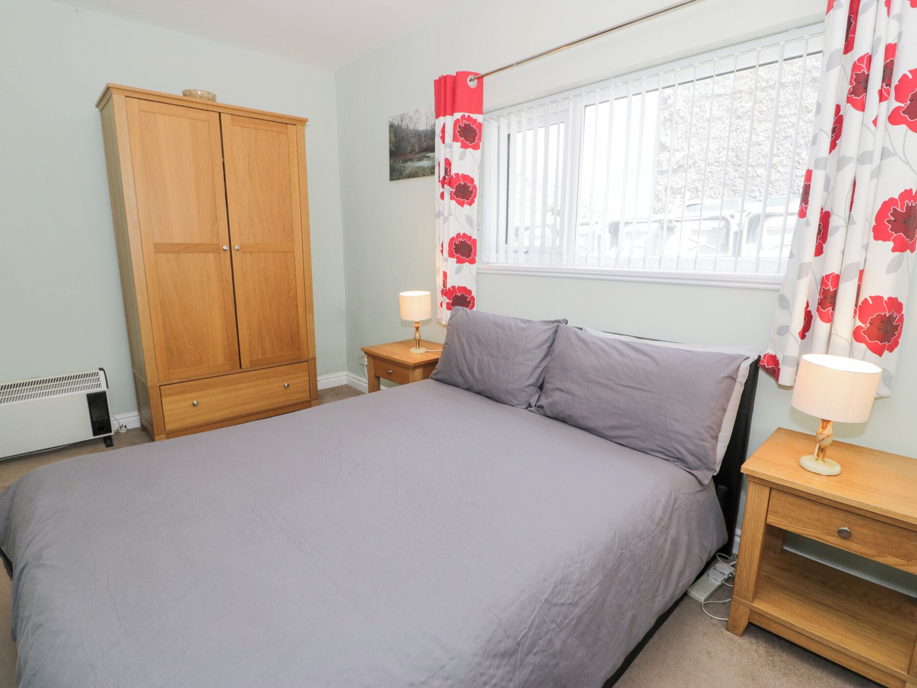 A bedroom with a large bed, a wooden wardrobe, two bedside tables with lamps, and a window with floral curtains.