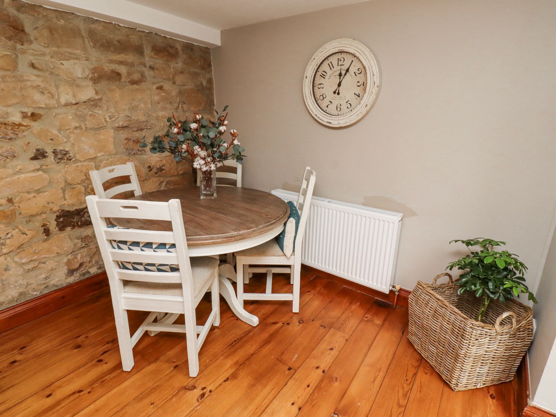 Un coin repas avec un mur en pierre, une table ronde en bois, des chaises blanches, une horloge, un radiateur et une plante dans un panier.