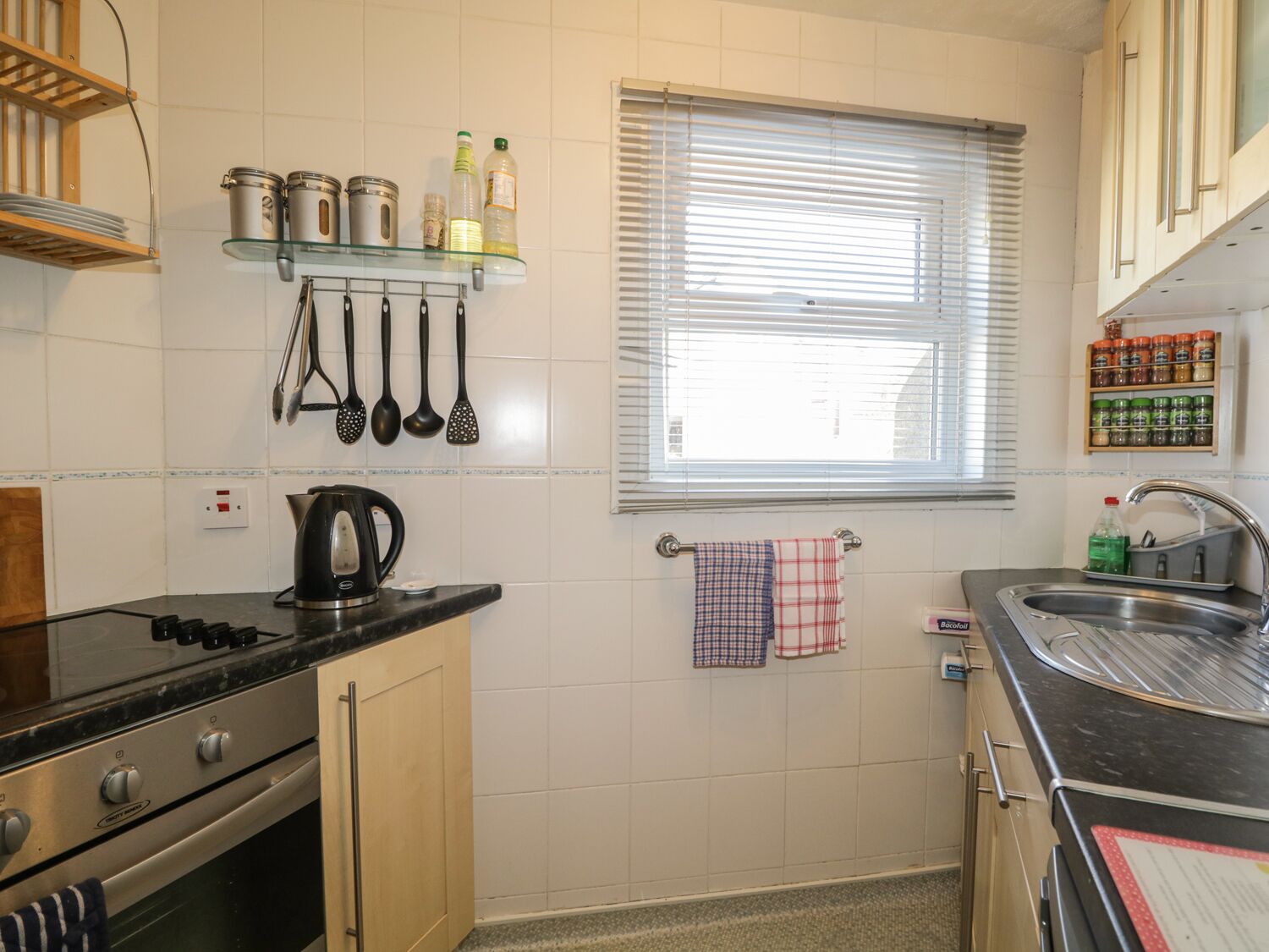 Une cuisine de taille réduite, équipée d’une cuisinière, d’une bouilloire et d’une fenêtre avec des stores.