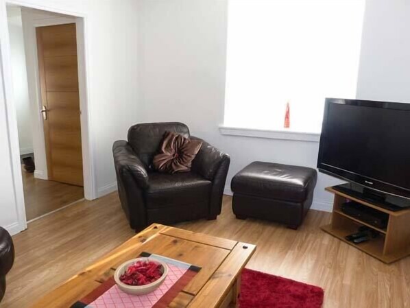 A living room with a leather sofa, a wooden coffee table with a bowl, a red rug, a television on a wooden stand, and a window with blinds. - Coastal Cottage (Fraserburgh)
