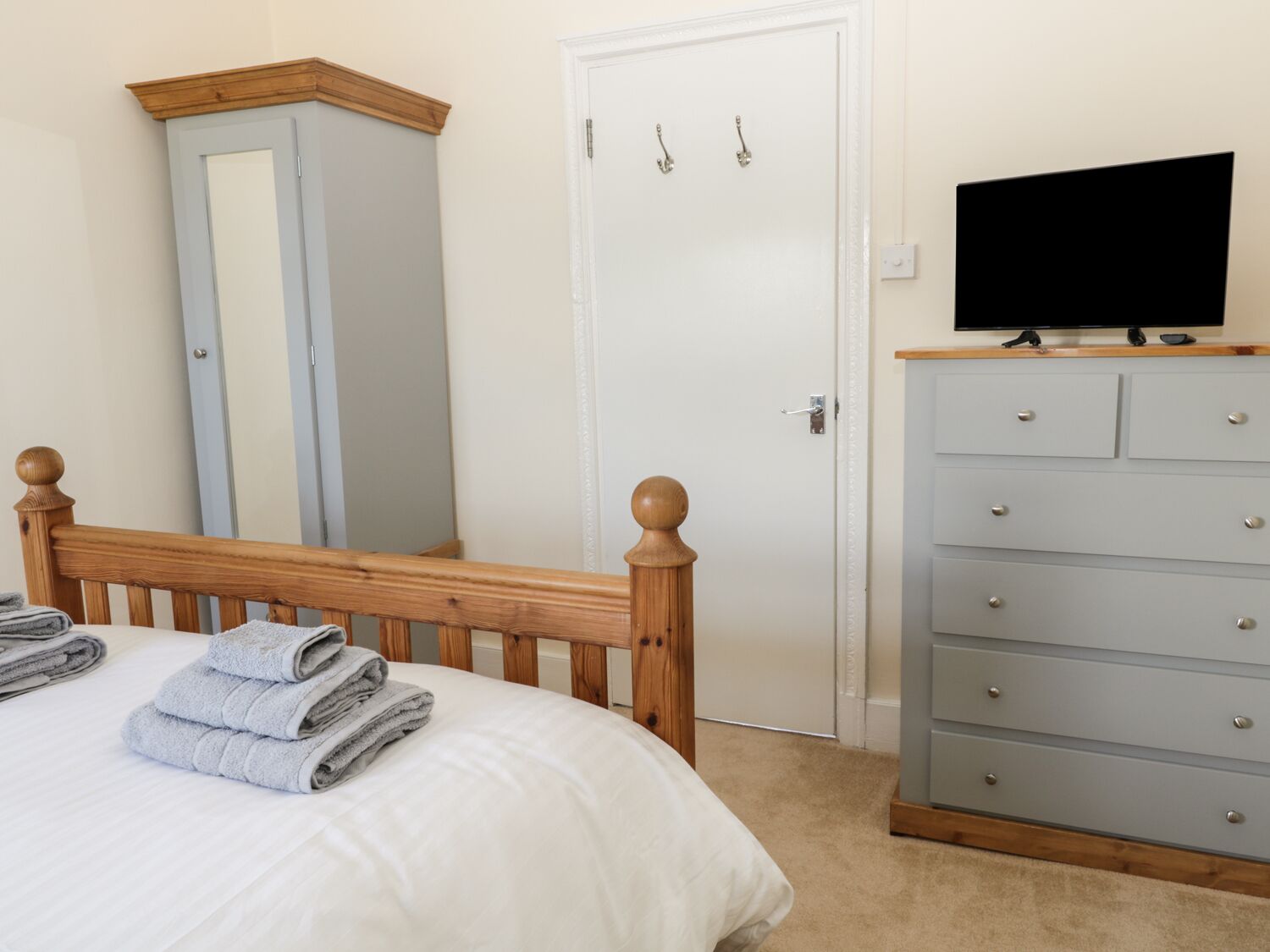 Une chambre avec un lit en bois, une commode grise, un téléviseur posé dessus et une porte blanche avec un miroir.