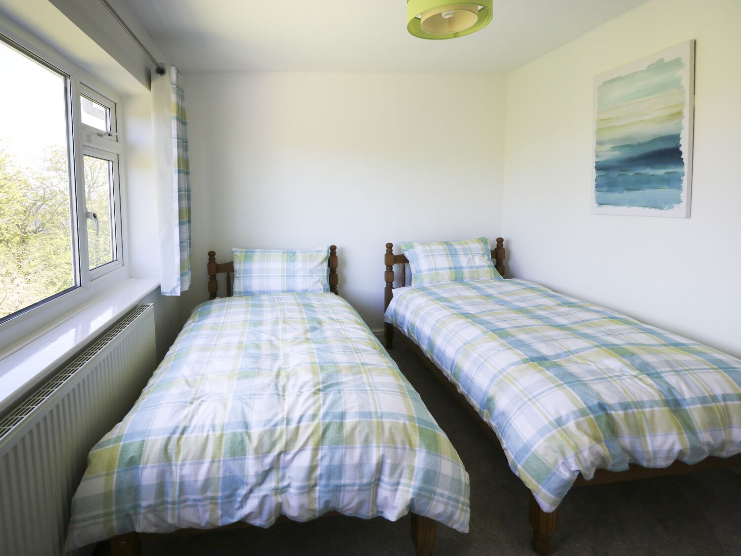 A room with two single beds, a radiator, a window with curtains, and a wall with a painting.