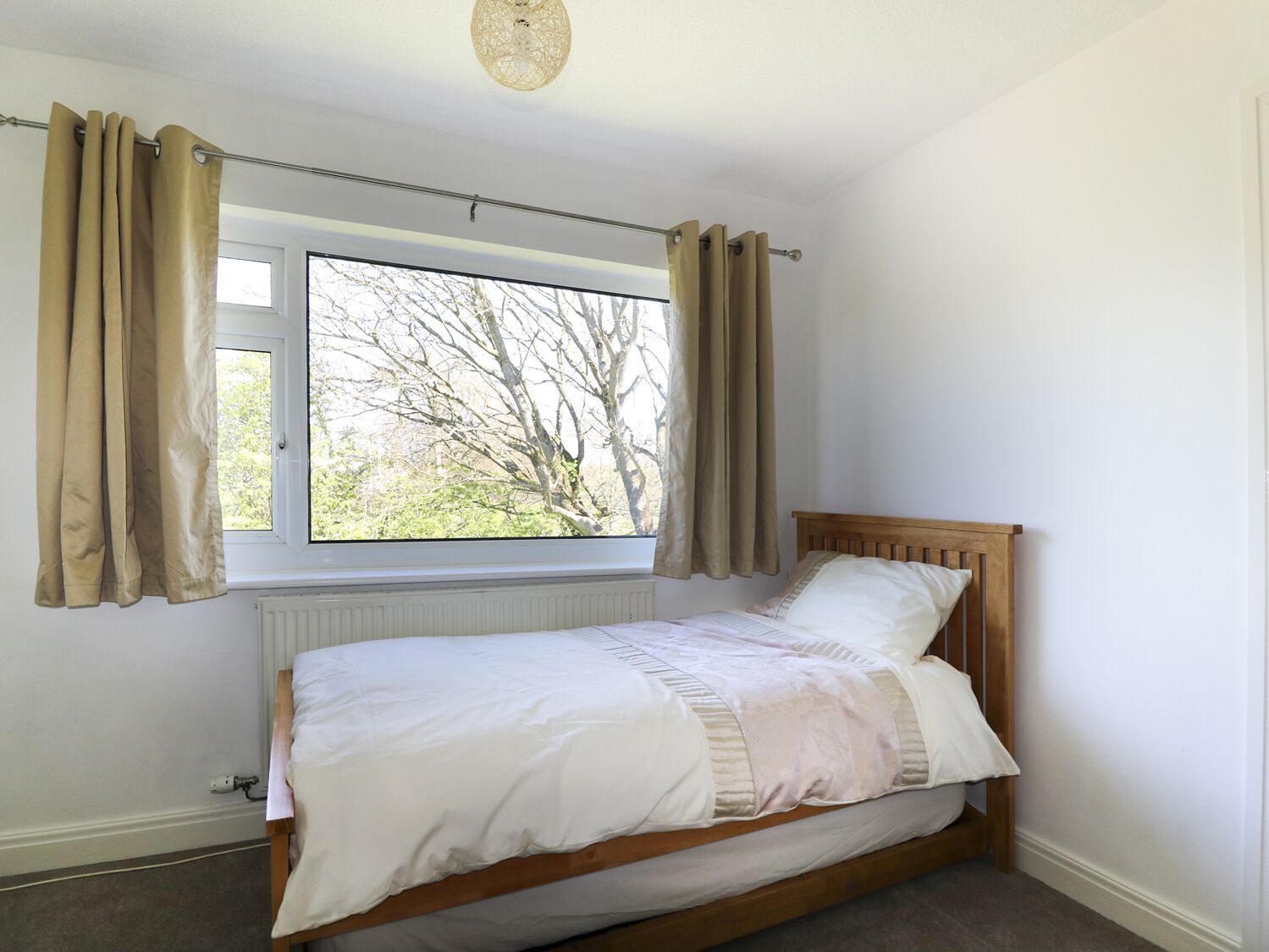 A bedroom with a wooden bed, beige curtains, a window with a view of trees, and a radiator.
