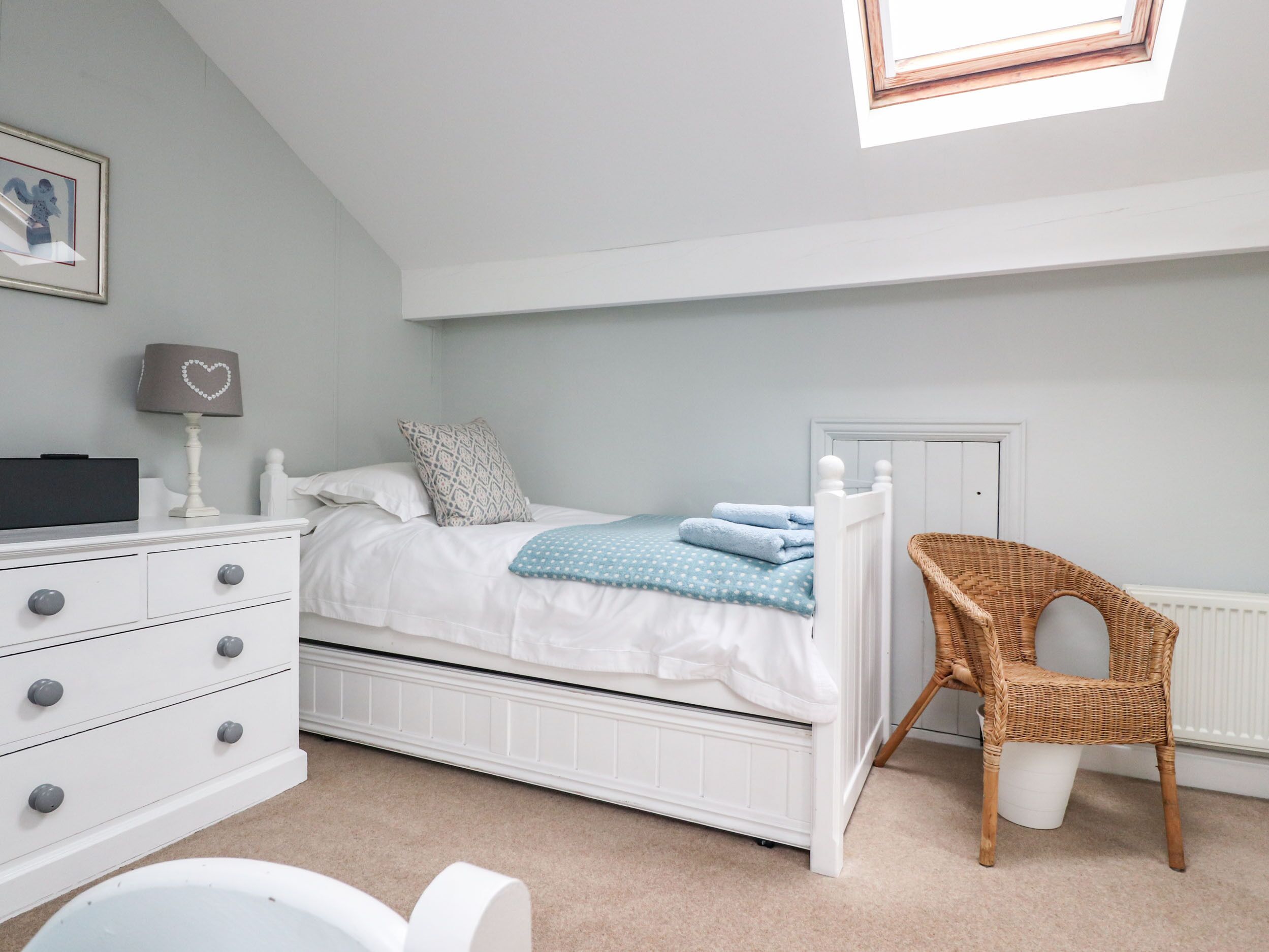 A bedroom with a white bed, a dresser with drawers, a wicker chair, and a skylight.