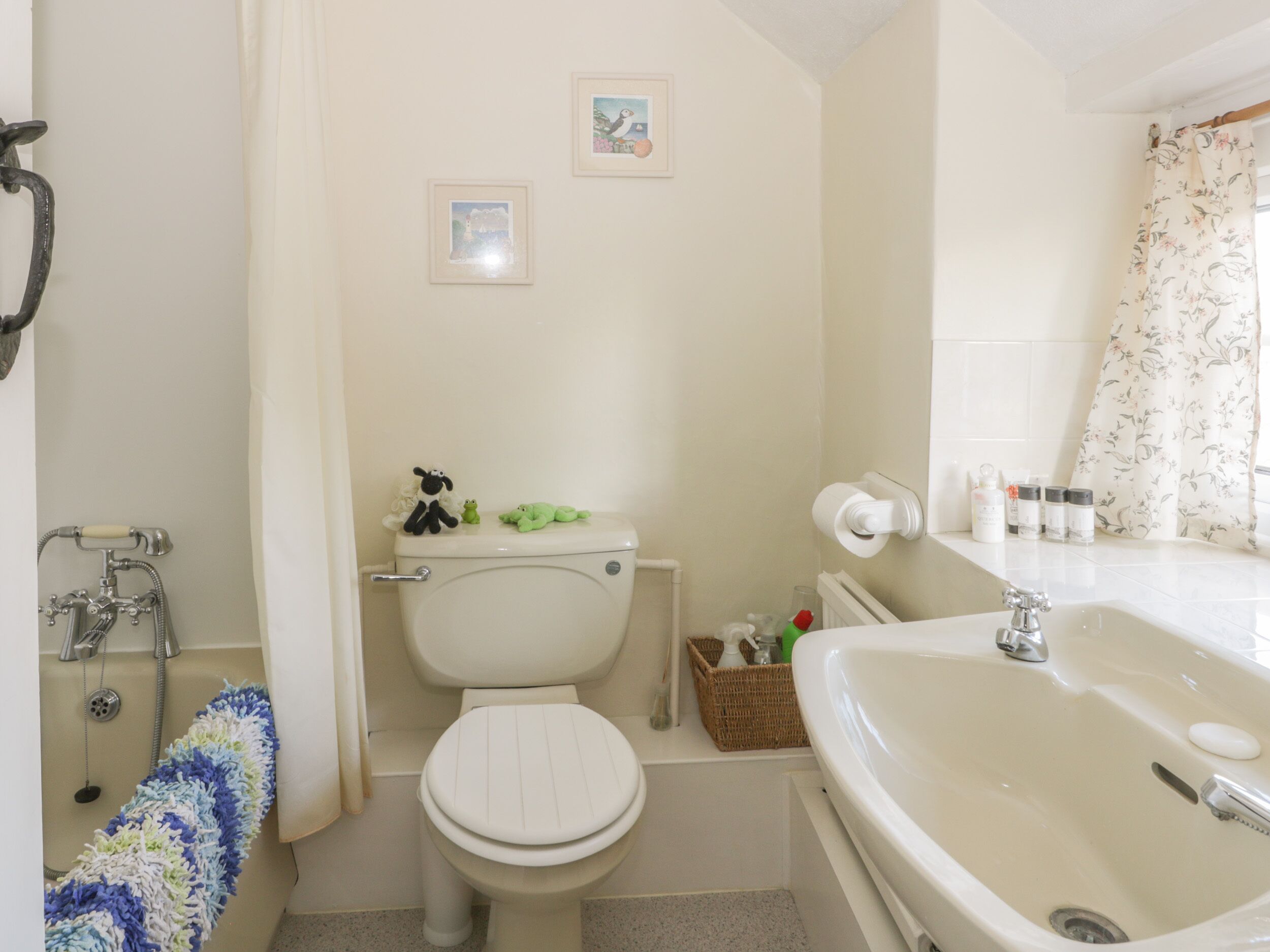 A bathroom with a toilet, bidet, and sink. There are two framed pictures on the wall, a basket with toiletries, and a shower curtain.
