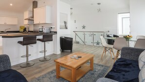 Ett modernt kök med vita skåp, en köksö och barstolar. Det finns en matplats med ett glasbord och stolar, samt ett soffbord i trä.