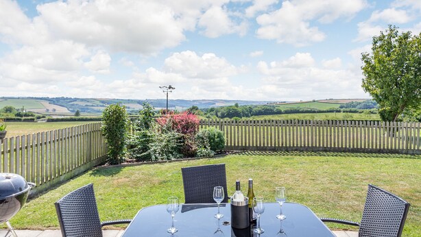 Un patio con una mesa preparada para dos personas, rodeado de sillas de mimbre, con vistas a un jardín cercado y colinas ondulantes.