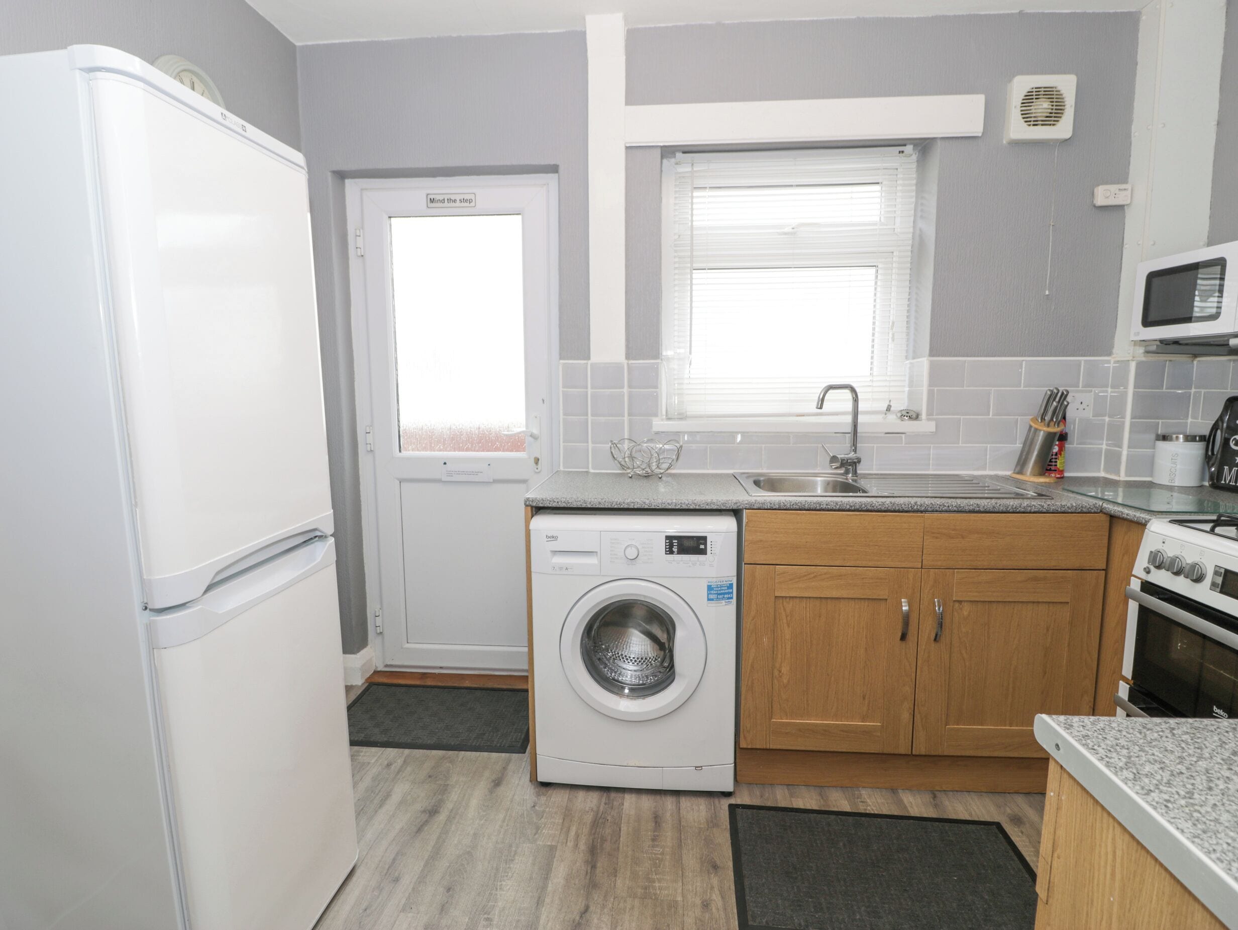 A compact kitchen with a washing machine, a refrigerator, a microwave, and wooden cabinets.