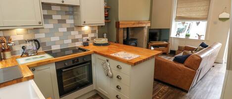 A compact kitchen with a wooden countertop, an oven, and a built-in stove. There is a small island with a kettle and a cutting board. The kitchen has light-colored cabinets and a tiled backsplash.