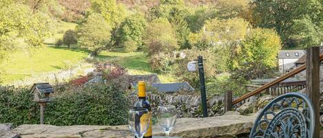 A stone table with a bottle and two glasses, overlooking a scenic valley with trees and a house.
