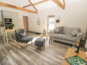 Een moderne woonkamer met een grijze bank, een grijze fauteuil en een houten salontafel. Er is een keukenhoek met een wit kastje en een oven. Een schaakbord staat op de salontafel.