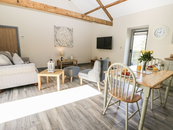 Een gezellige woonkamer met een bank, een eettafel met stoelen, een aan de muur bevestigde televisie en een houten plafond.