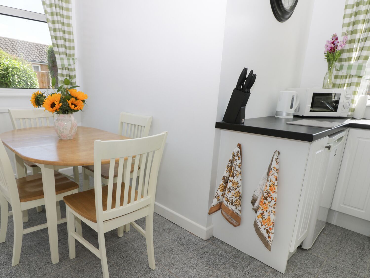 A kitchen with a wooden table and chairs, a microwave, a knife block, and a vase with flowers.