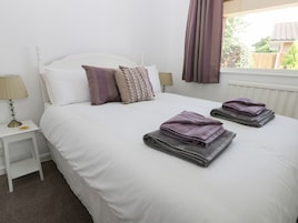 Una cama bien hecha con ropa blanca y dos toallas dobladas, una mesita de noche con lámpara, y una ventana con cortinas.