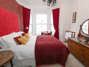 A bedroom with a large bed, red curtains, a wooden dresser, and a window with a view of buildings. - Abbeylands House (Llandudno)