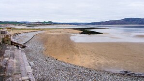 Una playa costera con piedras y arena, un promontorio rocoso y colinas a lo lejos.
