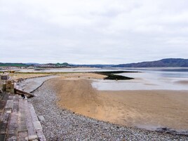 Una playa costera con piedras y arena, un promontorio rocoso y colinas a lo lejos.