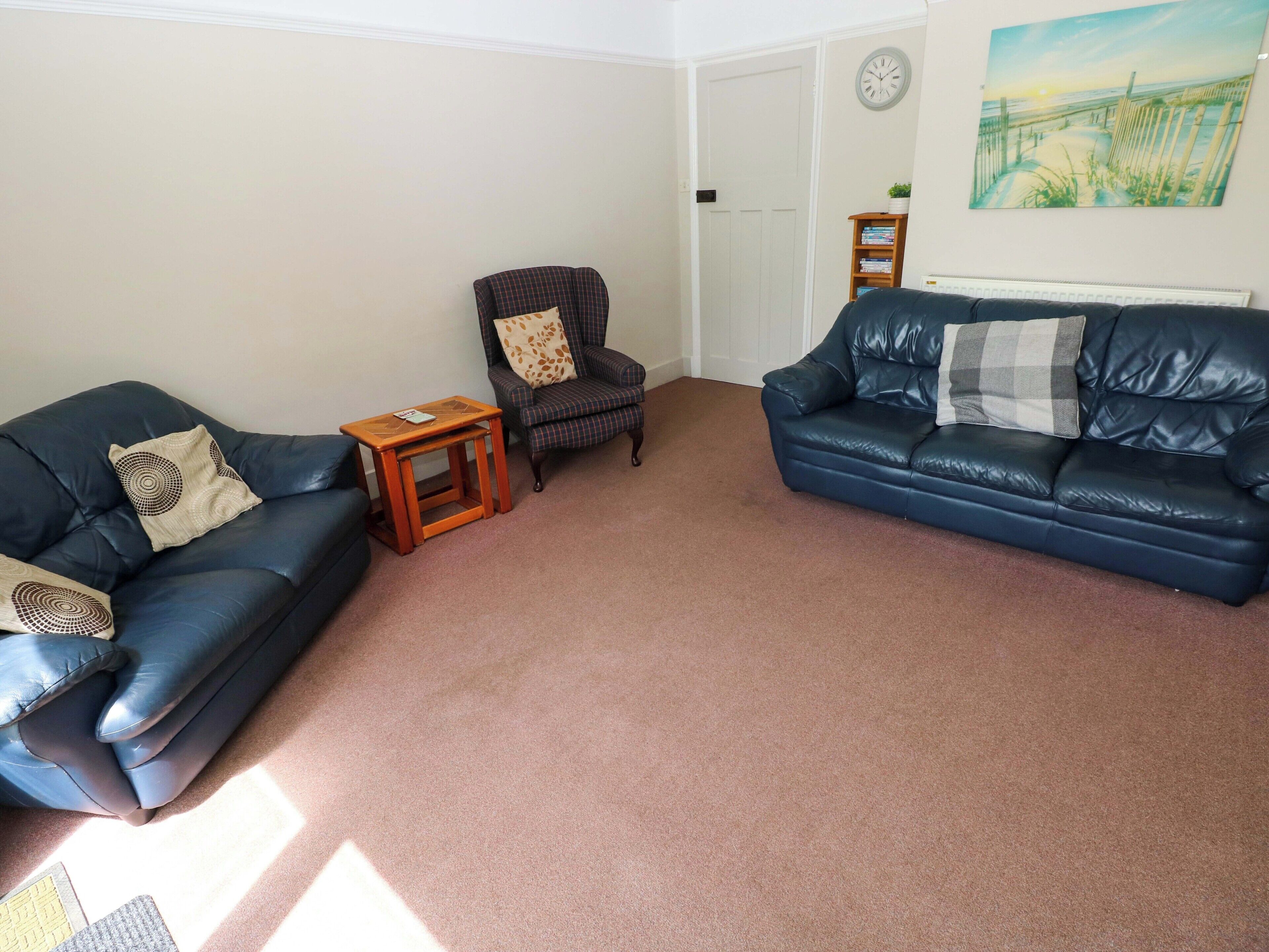 A living room with two dark blue leather sofas, a small wooden side table, a plaid armchair, and a painting on the wall.