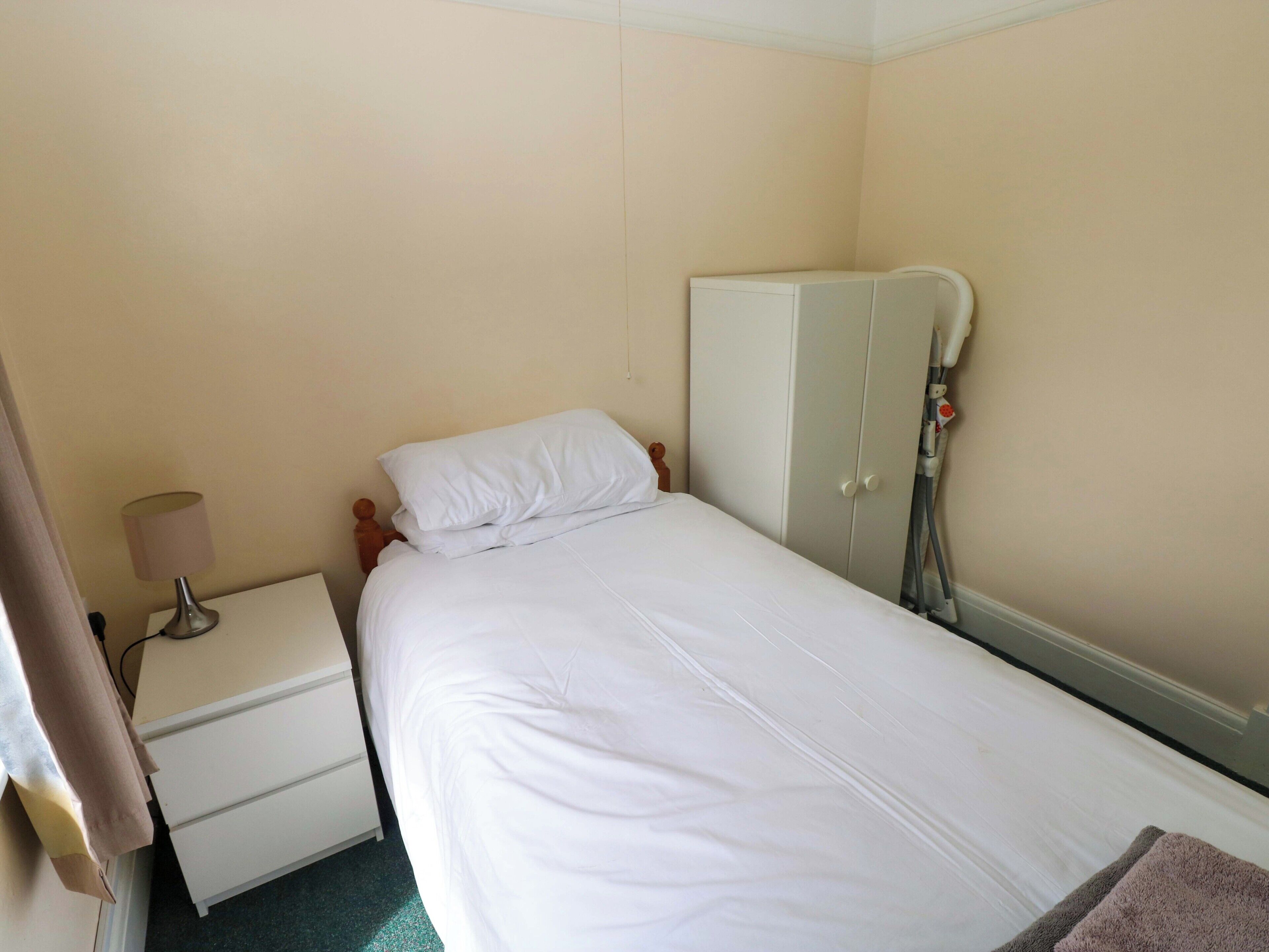A single bed with white bedding, a bedside table with a lamp, and a wardrobe.