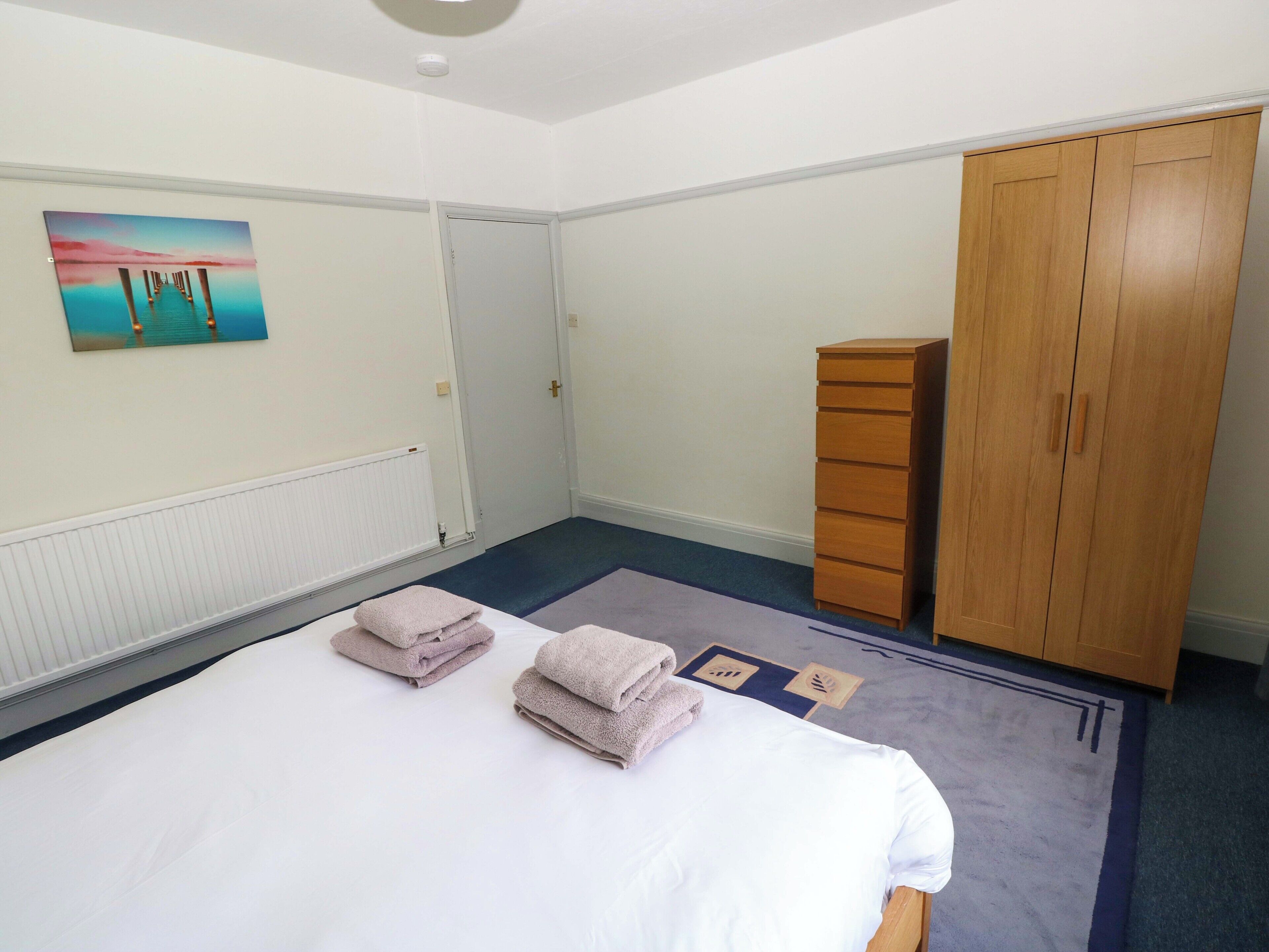 A bedroom with a bed, a wooden wardrobe, a radiator, a painting, and a carpet.