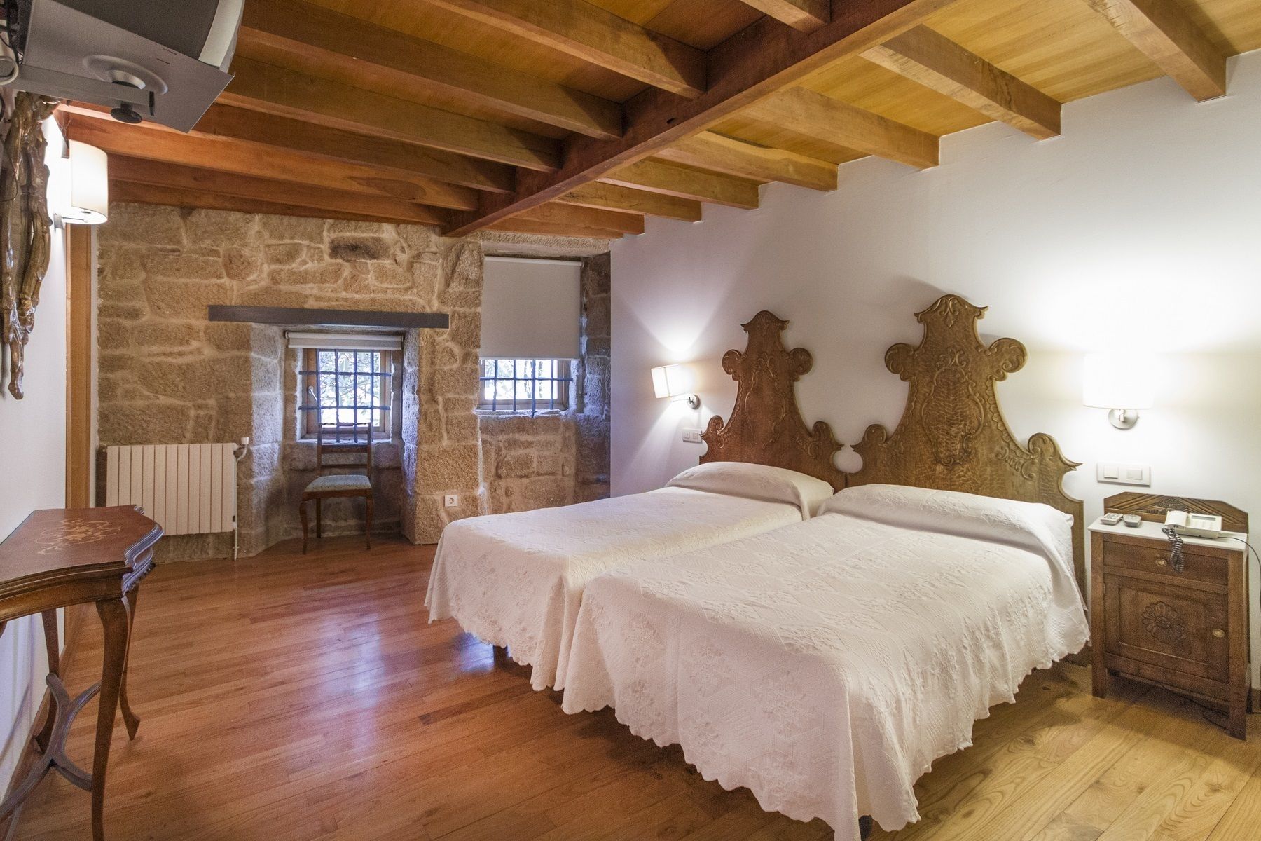 A bedroom with a stone wall, wooden beams, a double bed with white linens, a wooden nightstand, and a small table with a chair.