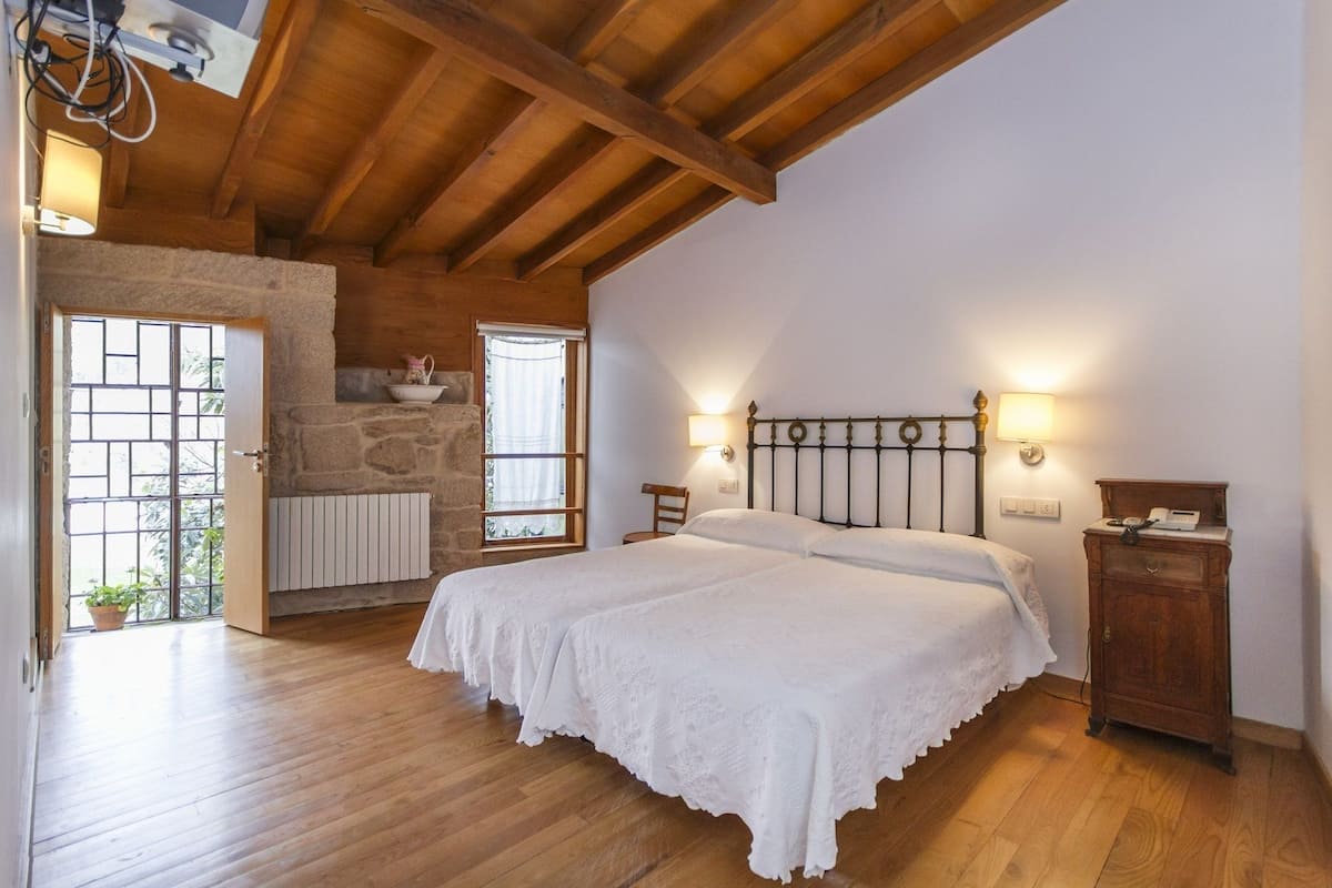 Un dormitorio con techo de madera, pared de piedra, cama con cubrecama blanco, dos lámparas de noche y mesita de noche de madera.