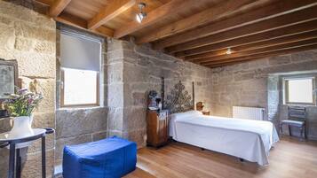 Un dormitorio rústico con pared de piedra, vigas de madera, una cama, una mesita con un florero y un banco azul.