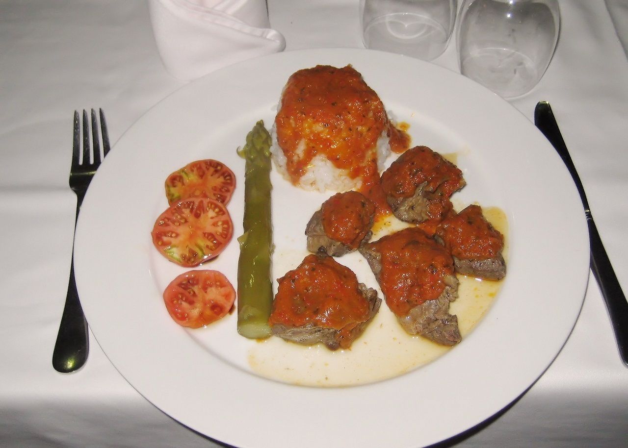 Un plato con pollo, tomates y un vegetal verde sobre un mantel blanco.