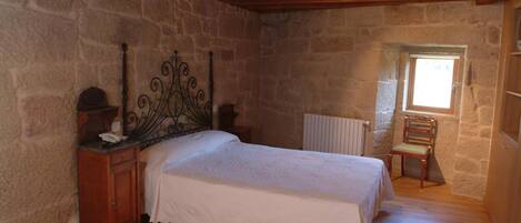A bedroom with a stone wall, a wooden bed, a nightstand, a chair, and a window.