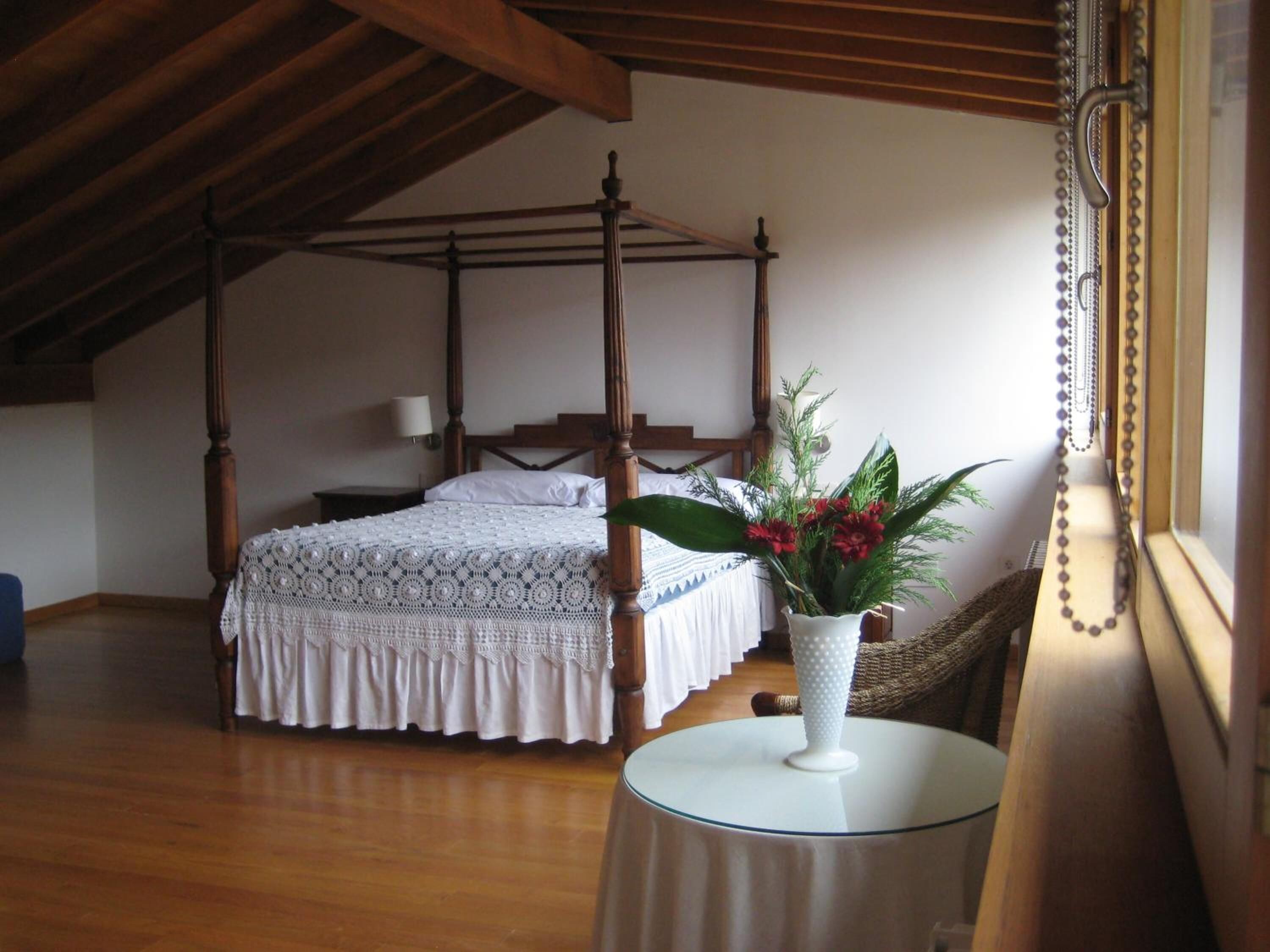 Un lit à baldaquin avec un couvre-lit blanc, une table de chevet ornée d’un vase de fleurs et une chaise en osier près d’une fenêtre avec un rideau.