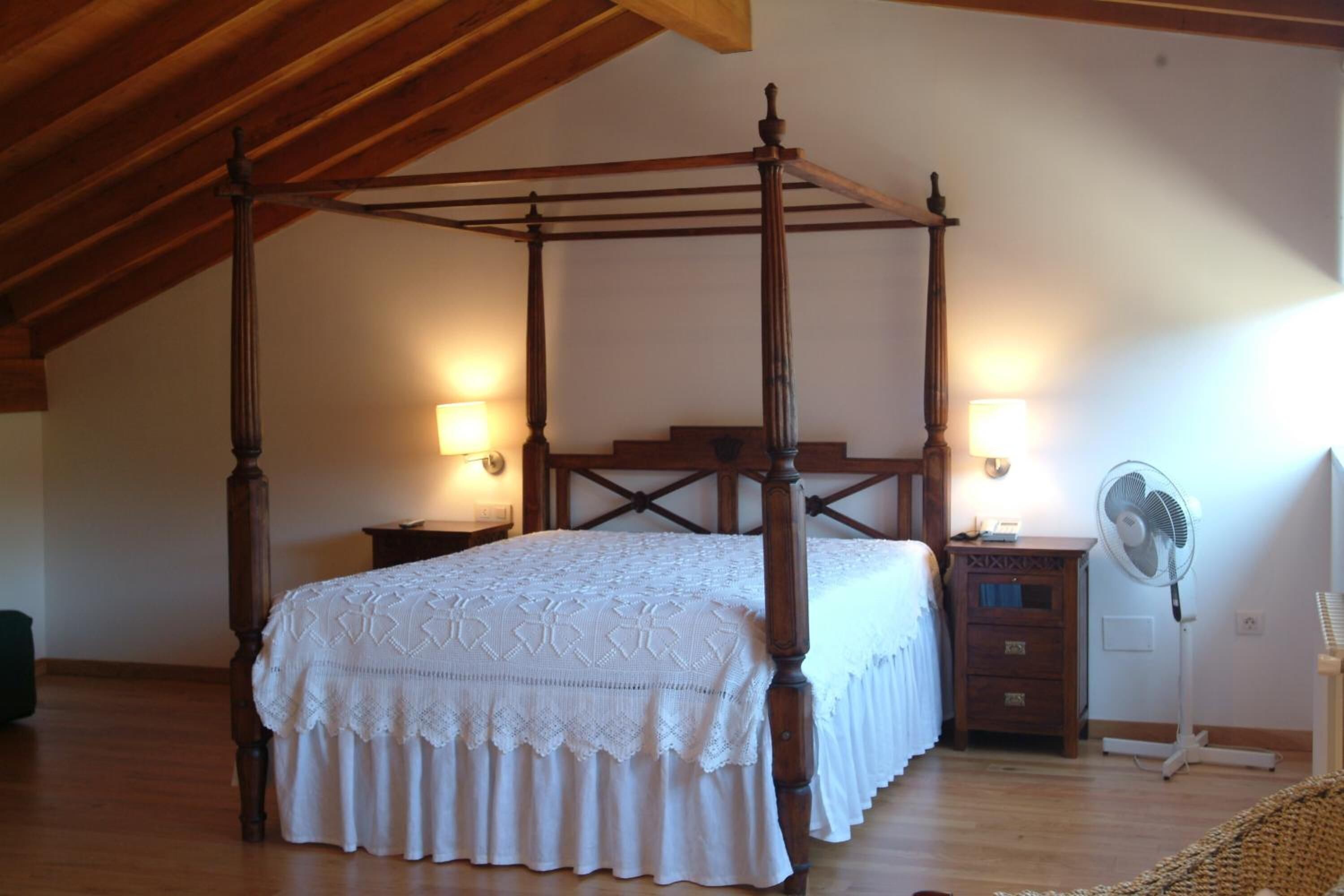 A four-poster bed with a white bedspread, two bedside lamps, a wooden nightstand, and a standing fan.