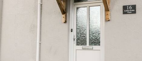 A white door with a frosted glass window, a wooden porch with a triangular roof, and a sign reading "16 Little Orme View".