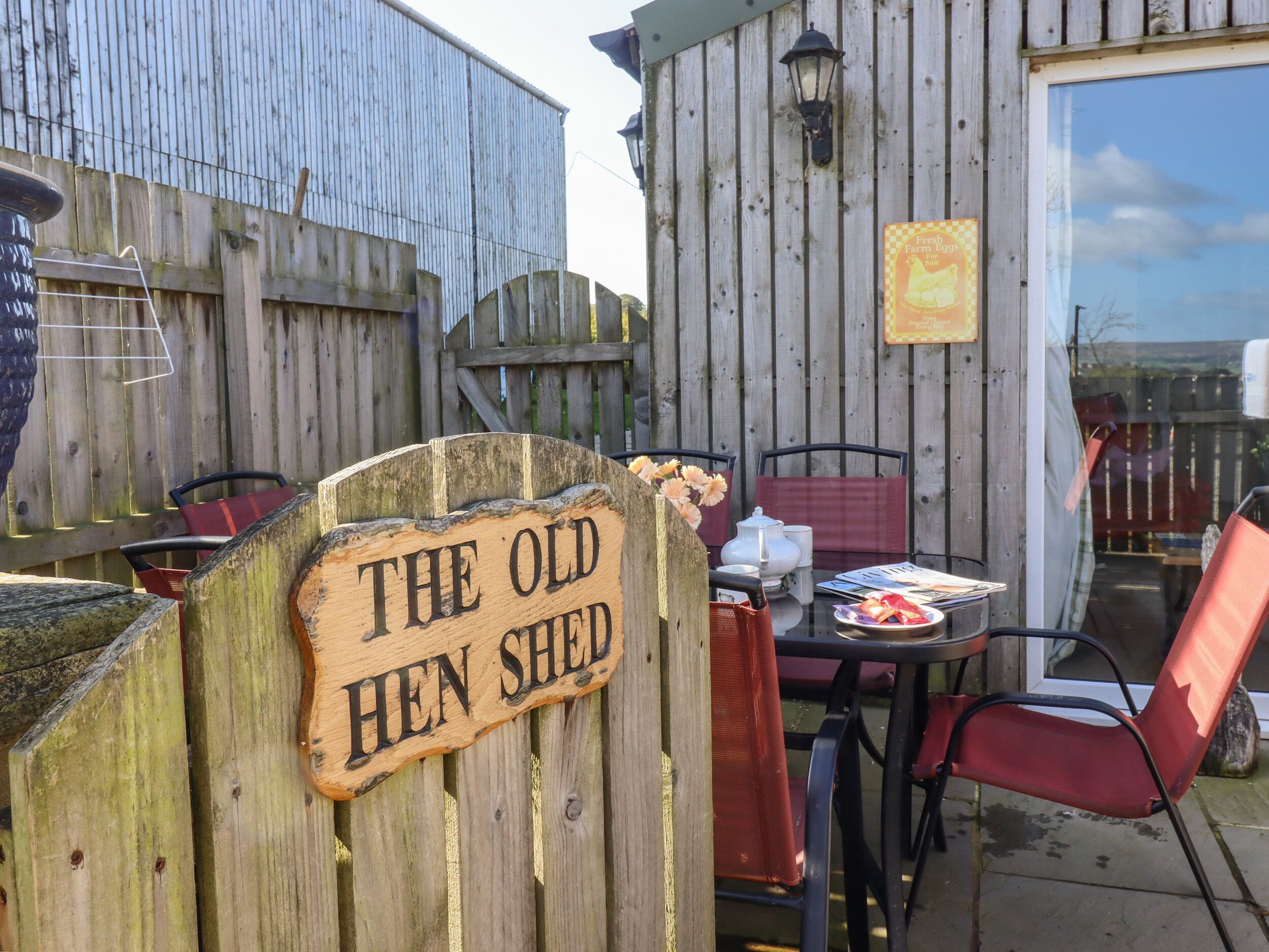 Ein Holzschild mit der Aufschrift 'THE OLD HEN SHED', ein rustikaler Außenbereich mit Tisch und Stühlen sowie ein Holzzaun im Hintergrund.