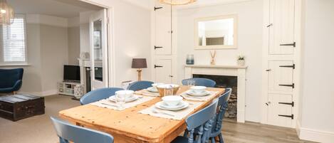 Un comedor con una mesa de madera preparada para cuatro personas, rodeada de sillas azules. Hay una lámpara colgante de mimbre sobre la mesa y una chimenea blanca con un espejo encima.