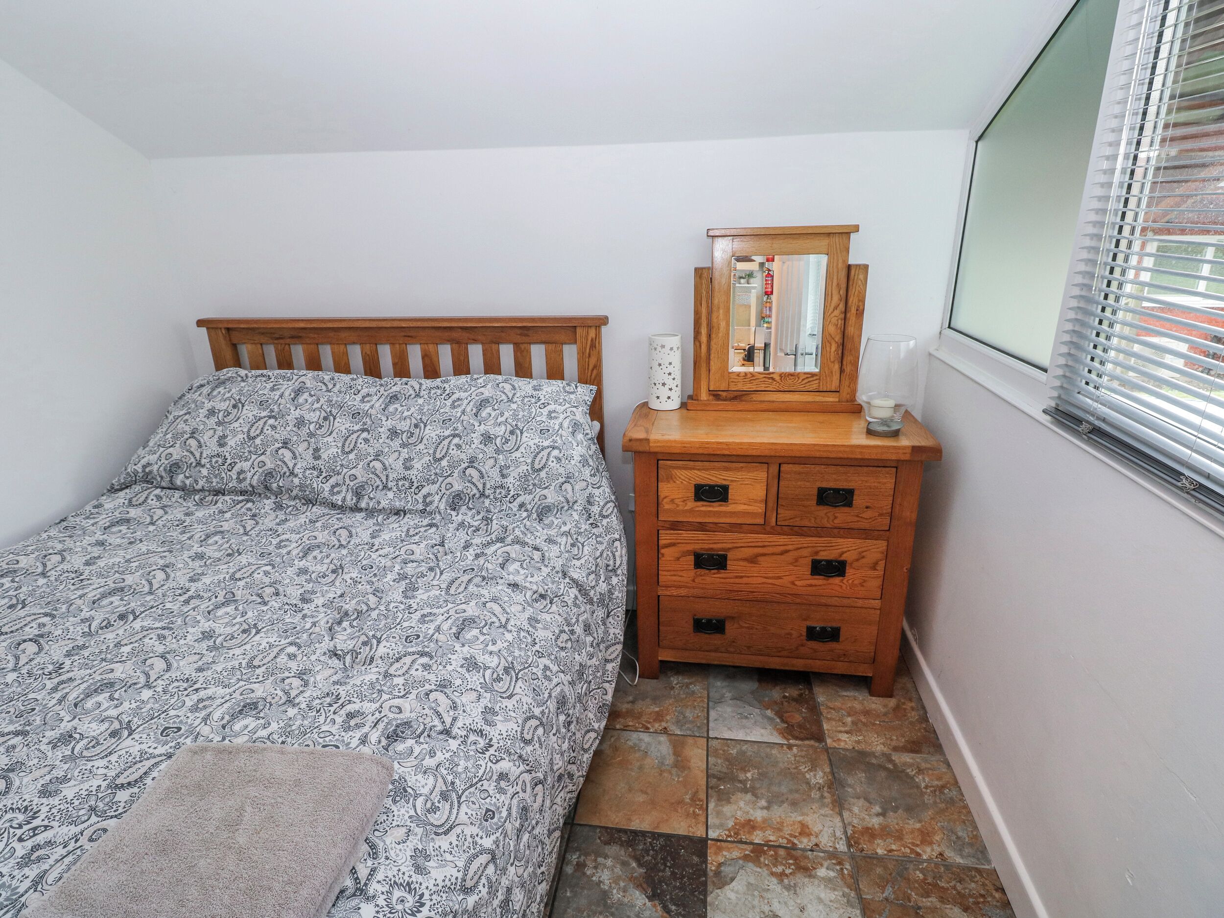 Een slaapkamer met een bed, een houten dressoir, een spiegel en een raam met jaloezieën.