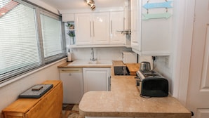 A compact kitchen with white cabinets, a built-in dishwasher, a toaster, and a window with blinds. - Trelessy Lodge (Narberth)