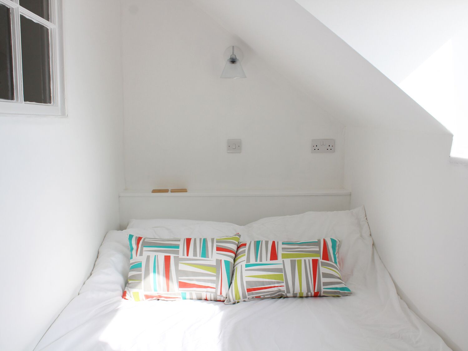 A small, white room with a sloped ceiling, a single bed with colorful pillows, and two electrical outlets on the wall.