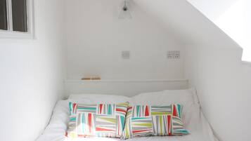 A small, white room with a sloped ceiling, a single bed with colorful pillows, and two electrical outlets on the wall.
