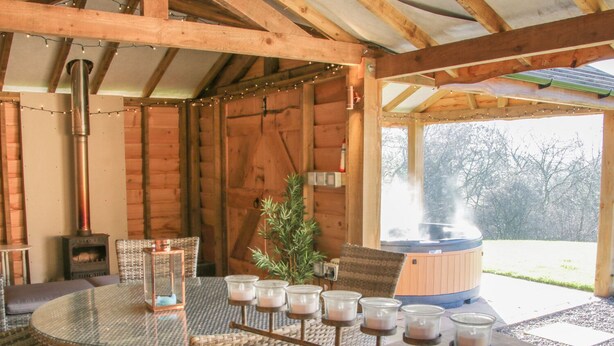 A wooden interior with a round glass table, wicker chairs, a small potted plant, a candle holder with multiple candles, a wood stove, and a hot tub outside. - Stoney-brook Lodge (Ludlow)
